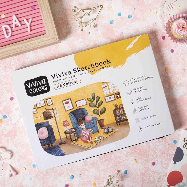 Peut inclure: Un carnet de croquis avec une couverture jaune et blanche illustrant une femme assise dans un fauteuil avec un chat. Le texte sur la couverture indique "Viviva Sketchbook Premium Handmade Sketchbooks A5 Cotton".