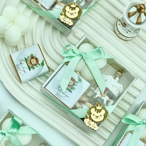 May include: Multiple gift boxes with clear lids, tied with pale green ribbons. Each box contains small white items, a decorative bottle, a toy horse, and a small square with a lion graphic and the text "Baby Lyn 2025".
