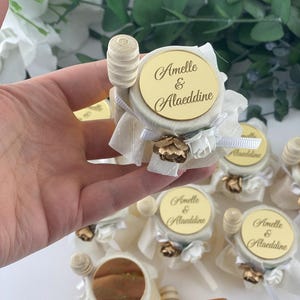 Könnte beinhalten: Kleine Honiggläser mit hölzernen Honiglöffeln. Jedes Glas ist mit einem goldfarbenen Deckel, einer weißen Stoffverpackung und einer goldfarbenen Blume verziert. Der Deckel trägt die Namen "Amelle & Alaeddine".