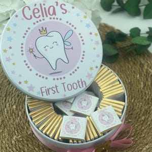 May include: A round, open tin filled with gold-wrapped chocolates. The lid features the text "Célia's" and "First Tooth" with a cartoon tooth illustration. The tin is decorated with pink, blue, and yellow stars.