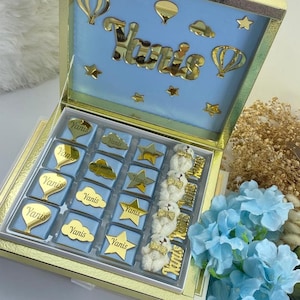 May include: A blue and gold gift box with a lid that has a blue background with gold stars, clouds, and hot air balloons. The lid also has the name "Yanis" written in gold letters. The box is filled with small blue and gold chocolate bars with the name "Yanis" on each one.
