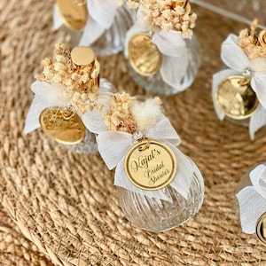 Several small, clear, ribbed glass perfume bottles with gold tops, white bows, dried flowers, and gold tags reading "Kajal's Bridal Shower" are arranged on a woven mat.