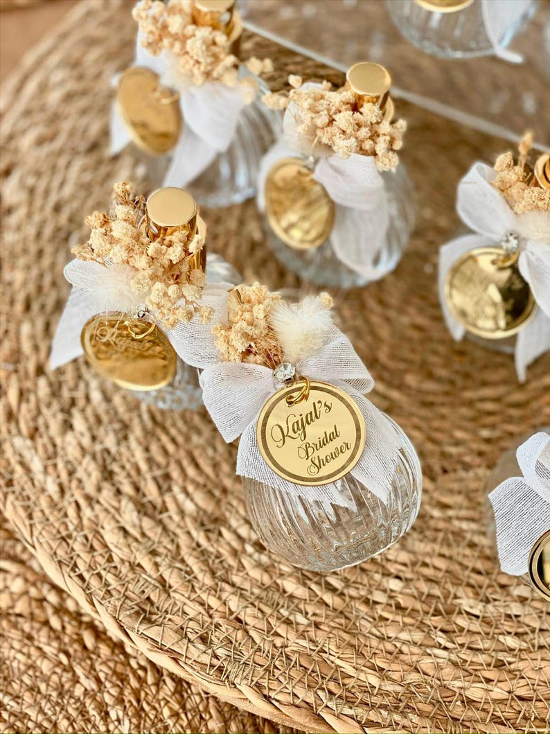 Several small, clear, ribbed glass perfume bottles with gold tops, white bows, dried flowers, and gold tags reading "Kajal's Bridal Shower" are arranged on a woven mat.