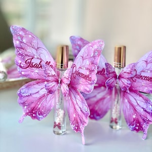 May include: Pink butterfly-shaped decorations with glitter accents and the text "Isabella Quincea&ntilde;era" on the wings. The decorations are paired with small perfume bottles with gold caps.