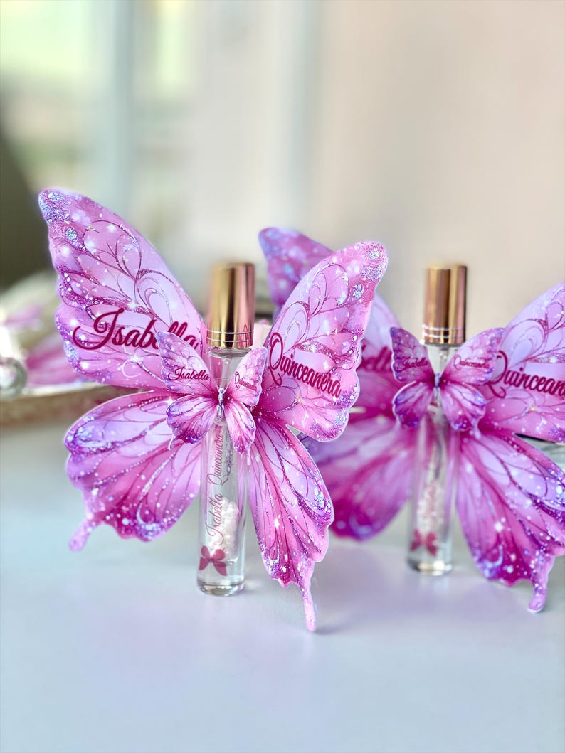 May include: Pink butterfly-shaped decorations with glitter accents and the text "Isabella Quincea&ntilde;era" on the wings. The decorations are paired with small perfume bottles with gold caps.