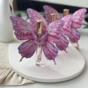 May include: Clear glass perfume bottles with gold caps, adorned with pink and purple butterfly wings. The wings have glitter accents and the name "Victoria" is written on the wings. The bottles are displayed on a white, round platform.