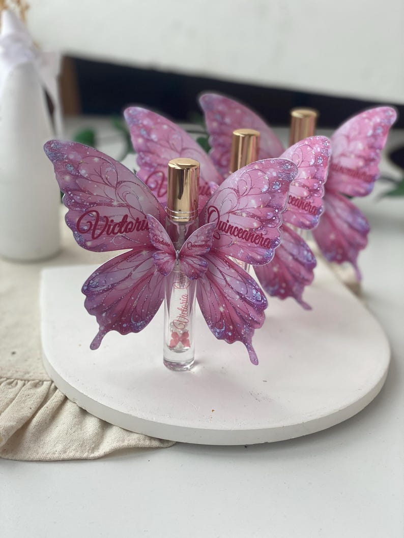 May include: Clear glass perfume bottles with gold caps, adorned with pink and purple butterfly wings. The wings have glitter accents and the name "Victoria" is written on the wings. The bottles are displayed on a white, round platform.