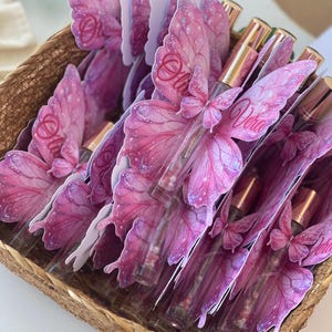 May include: A woven basket filled with perfume bottles. Each bottle is adorned with a pink butterfly wing design, with the name "Olivia" or "Vera" printed on the wings. The bottles have gold-colored caps.