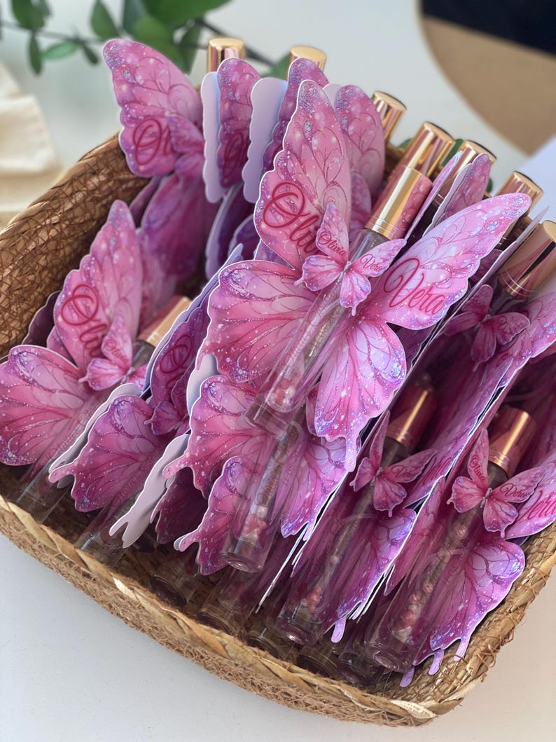 May include: A woven basket filled with perfume bottles. Each bottle is adorned with a pink butterfly wing design, with the name "Olivia" or "Vera" printed on the wings. The bottles have gold-colored caps.
