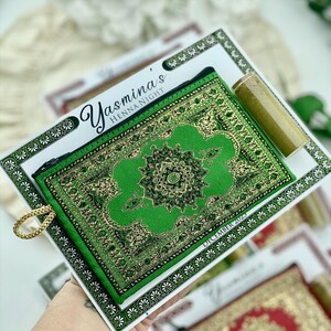 A hand holds up a green patterned mini wallet favor set, with a henna tube and a personalized card visible.