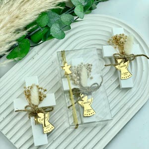 May include: Three white wooden crosses decorated with dried flowers and a gold angel charm with the name Camilla and the date of baptism.