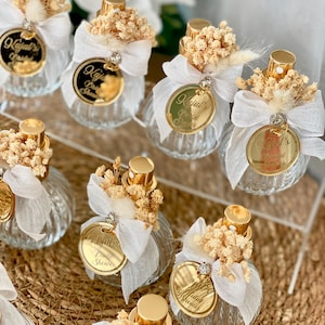 Eight small, clear, ribbed glass perfume bottles with gold tops, white bows, dried flowers, and gold tags are arranged on a woven mat and clear stand.