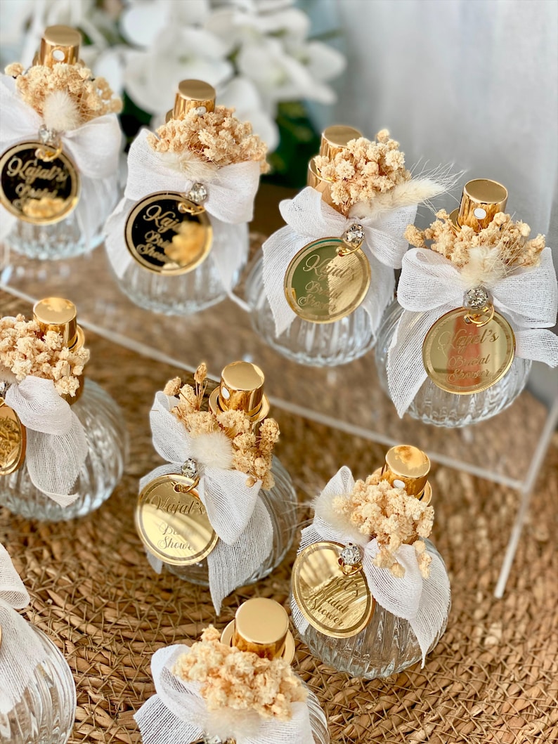 Eight small, clear, ribbed glass perfume bottles with gold tops, white bows, dried flowers, and gold tags are arranged on a woven mat and clear stand.