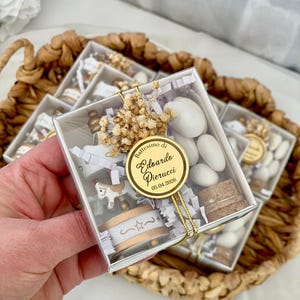 May include: Clear square favor boxes filled with white sugared almonds, a miniature carousel horse, and a cork. A gold sticker with text is attached to the box. The boxes are in a woven basket.