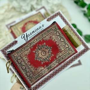 A hand holds a red patterned mini wallet favor set, which includes a henna tube and a personalized card.