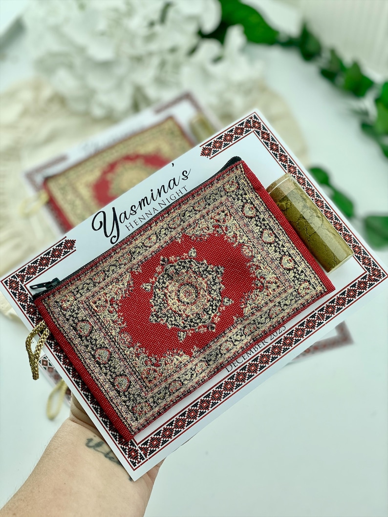 A hand holds a red patterned mini wallet favor set, which includes a henna tube and a personalized card.