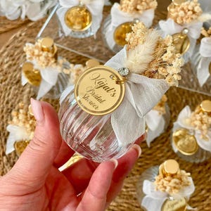 A hand holds a small, clear, ribbed glass perfume bottle with a gold top, light gray bow, dried flowers, and a gold tag reading "Kajal's Bridal Shower."