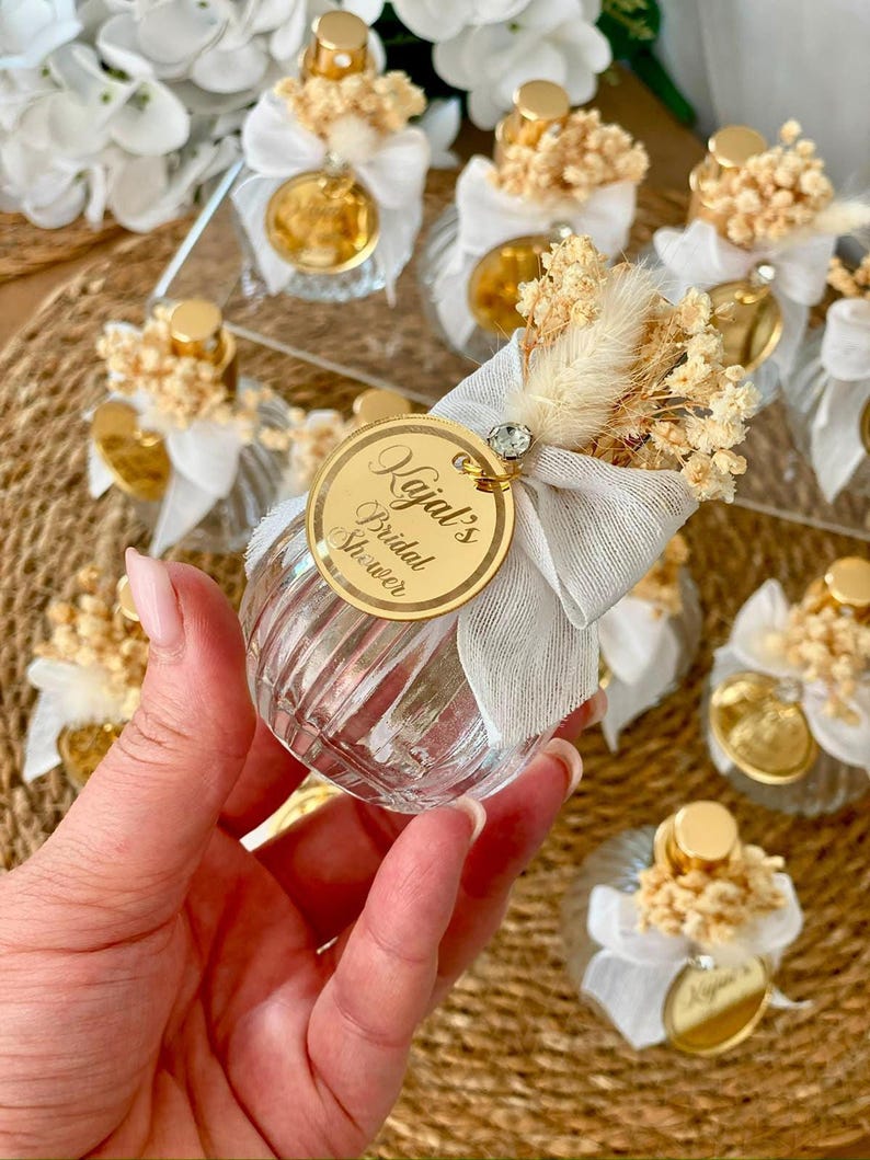 A hand holds a small, clear, ribbed glass perfume bottle with a gold top, light gray bow, dried flowers, and a gold tag reading "Kajal's Bridal Shower."