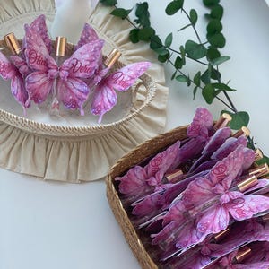 May include: Several perfume bottles with butterfly-shaped labels in a woven basket. The labels are pink and purple with the name "Olivia Vera" printed on them. The bottles have gold-colored caps. A second basket contains more bottles. The background is white.