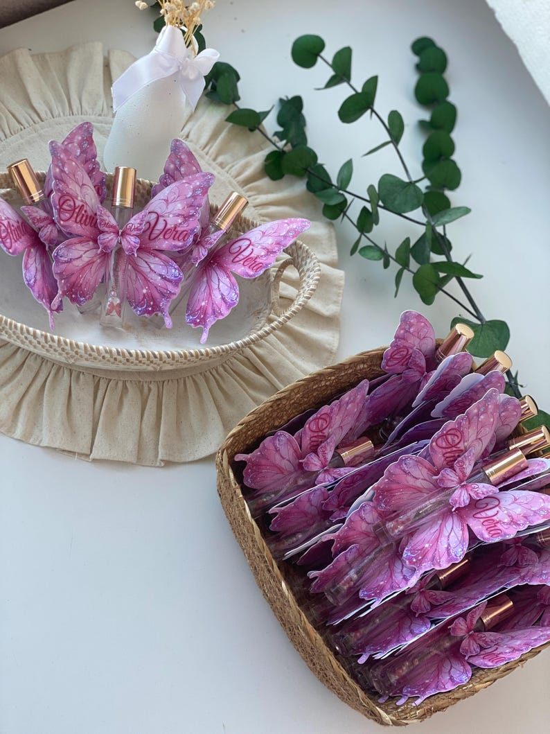May include: Several perfume bottles with butterfly-shaped labels in a woven basket. The labels are pink and purple with the name "Olivia Vera" printed on them. The bottles have gold-colored caps. A second basket contains more bottles. The background is white.