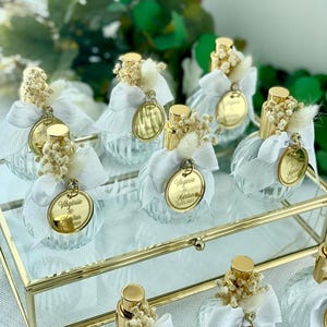 Nine small, clear glass perfume bottles with a ridged design, each adorned with a white bow, dried flowers, and a gold-rimmed tag, are arranged on a gold-framed glass tray.
