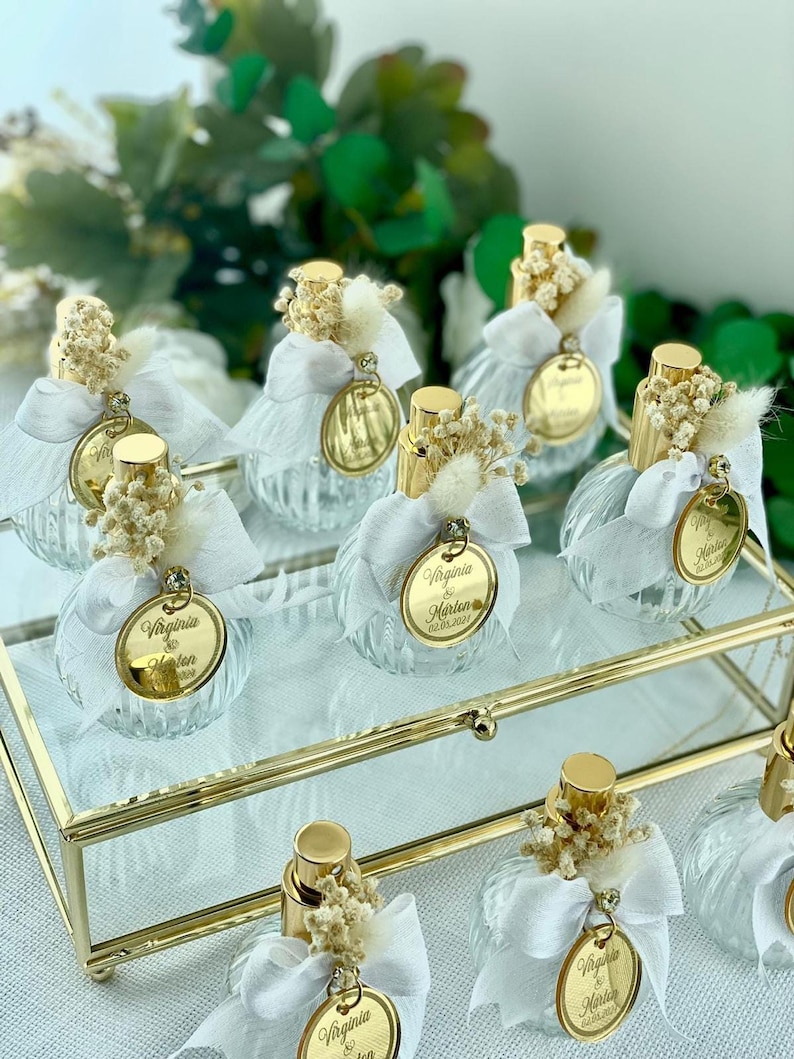 Nine small, clear glass perfume bottles with a ridged design, each adorned with a white bow, dried flowers, and a gold-rimmed tag, are arranged on a gold-framed glass tray.