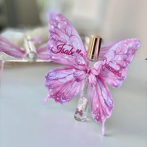 May include: A clear glass perfume bottle with a gold cap, adorned with pink butterfly wings. The wings are decorated with glitter and the name "Isabella Quinceanera". The bottle has a pink bow.