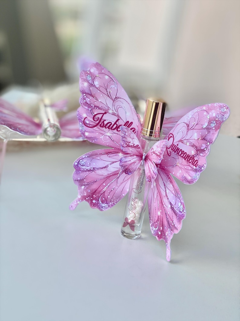 May include: A clear glass perfume bottle with a gold cap, adorned with pink butterfly wings. The wings are decorated with glitter and the name "Isabella Quinceanera". The bottle has a pink bow.