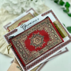 A hand holds a red patterned mini wallet favor set, which includes a henna tube and a personalized card.