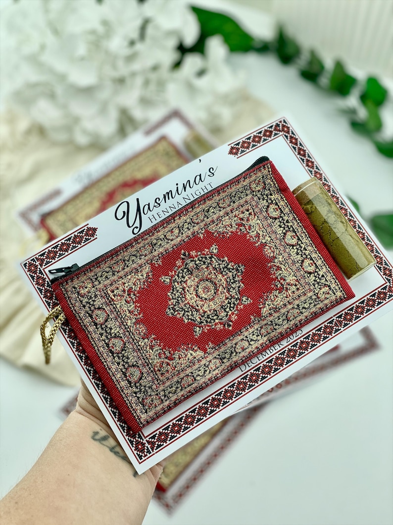 A hand holds a red patterned mini wallet favor set, which includes a henna tube and a personalized card.
