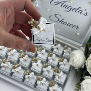 May include: A white box with a white bow and pearl embellishment. The box has the text "Angela's Shower" printed on it. The box is part of a larger set of similar boxes, all arranged in a white tray.