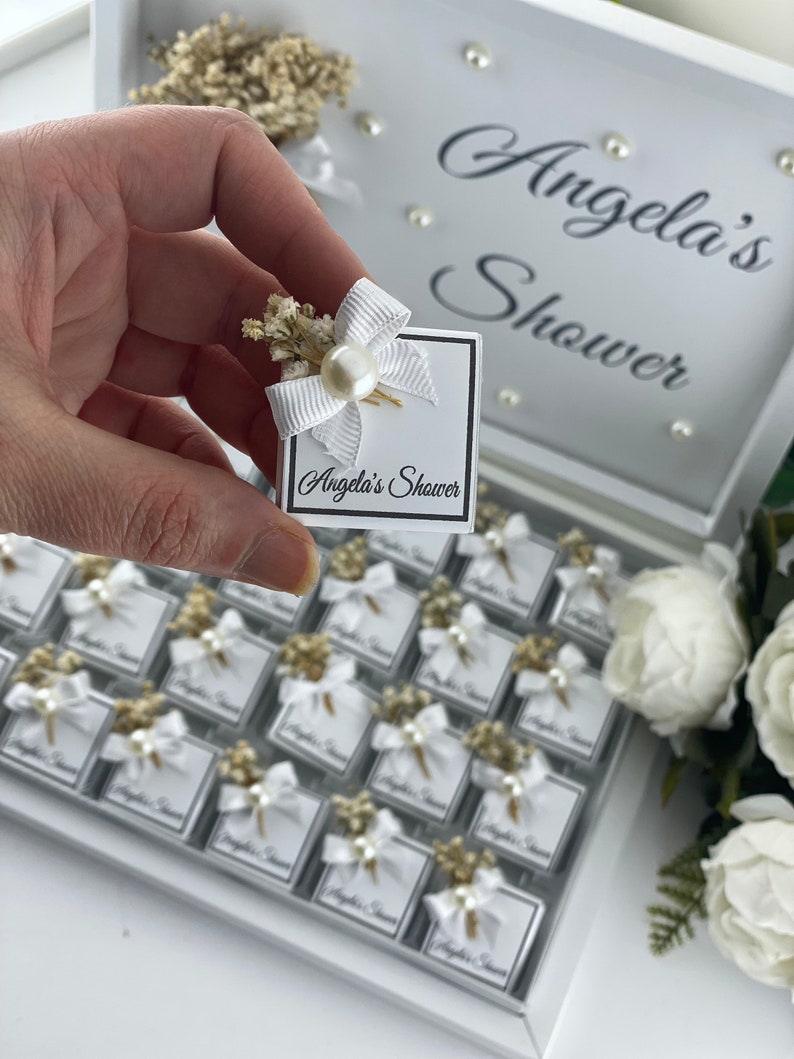 May include: A white box with a white bow and pearl embellishment. The box has the text "Angela's Shower" printed on it. The box is part of a larger set of similar boxes, all arranged in a white tray.
