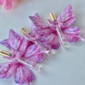May include: Three butterfly-shaped favors with clear tubes and gold caps. The pink and purple butterfly wings have the name "Isabella" and the word "Quinceanera" printed on them. The favors are on a white surface.
