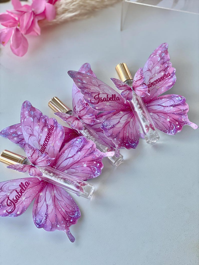 May include: Three butterfly-shaped favors with clear tubes and gold caps. The pink and purple butterfly wings have the name "Isabella" and the word "Quinceanera" printed on them. The favors are on a white surface.