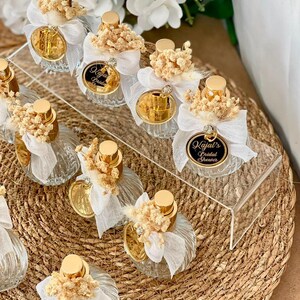 Eight small, clear, ribbed glass perfume bottles with gold tops, white bows, dried flowers, and gold tags are arranged on a woven mat and clear stand.