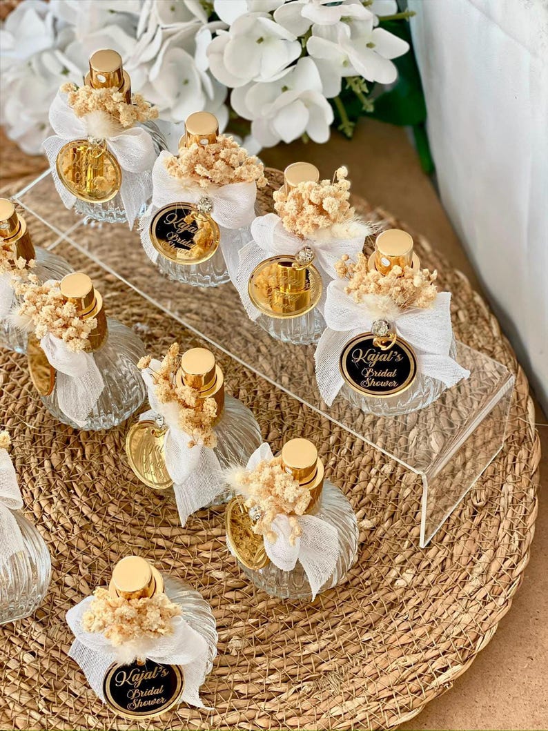Eight small, clear, ribbed glass perfume bottles with gold tops, white bows, dried flowers, and gold tags are arranged on a woven mat and clear stand.