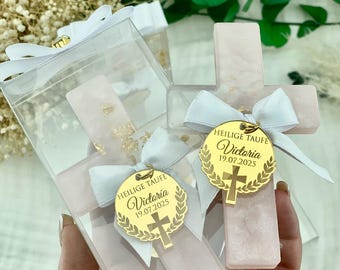 May include: Pink cross-shaped favors with gold accents, presented in clear boxes tied with white ribbons. Each favor has a gold tag that reads "HEILIGE TAUFE Victoria 19.07.2025" and a cross design. The favors are displayed against a backdrop of greenery.