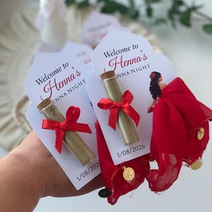 Puede incluir: Recuerdos de la Noche de Henna. Pequeñas botellas de vidrio con tapones de corcho, llenas de un polvo verde, atadas con cintas rojas y unidas a tarjetas blancas. Las tarjetas dicen "Bienvenida a la Noche de Henna" e incluyen una fecha. Tela roja con monedas de oro adjunta.