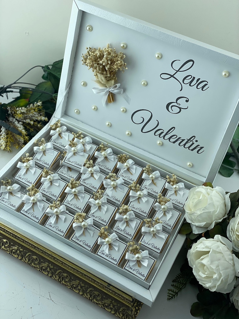 May include: White gift box with a gold trim, filled with small white boxes with a white bow and a small bouquet of dried flowers. Each box has the text "Leva & Valentin" printed on it.