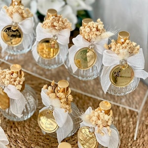 Eight small, clear, ribbed glass perfume bottles with gold tops, white bows, dried flowers, and gold tags are arranged on a woven mat and clear stand.