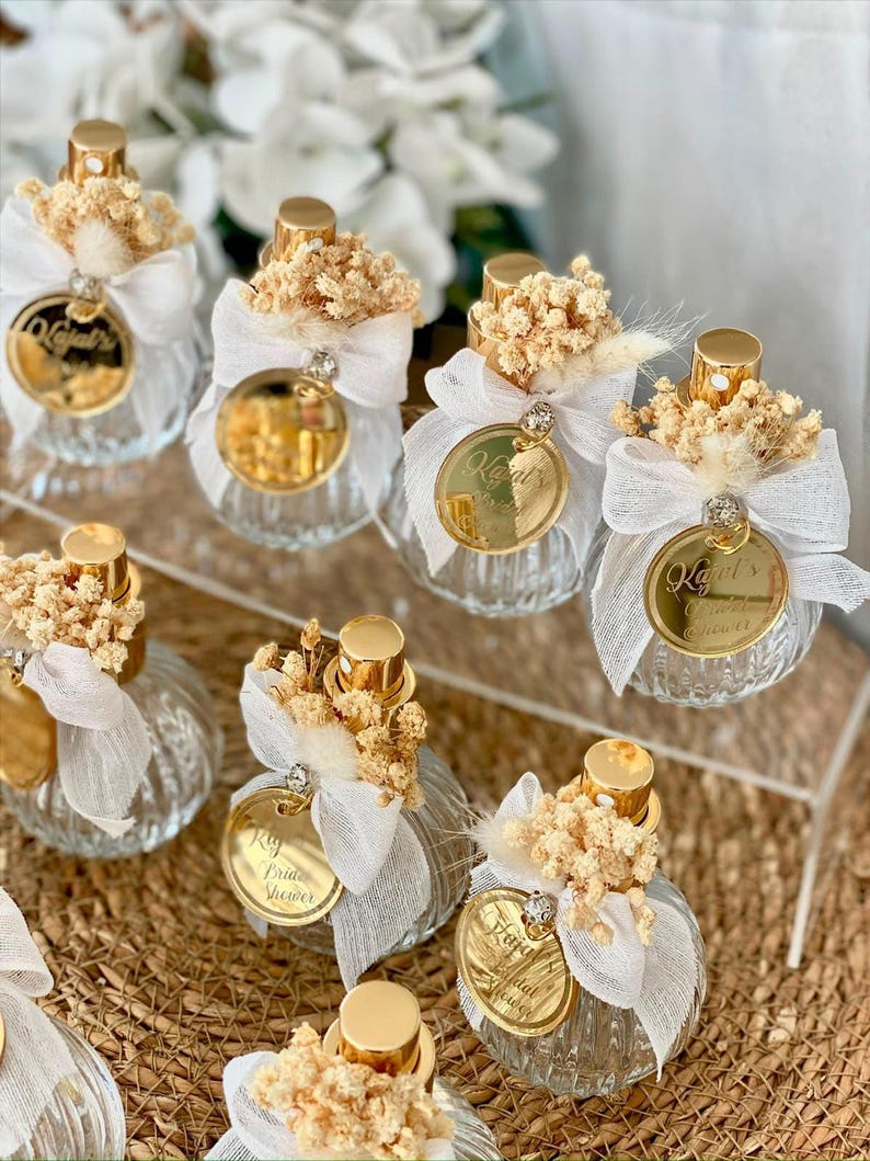 Eight small, clear, ribbed glass perfume bottles with gold tops, white bows, dried flowers, and gold tags are arranged on a woven mat and clear stand.