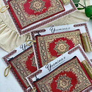 Four red patterned mini wallet favor sets are arranged on a white surface, each with a henna tube and a personalized card.