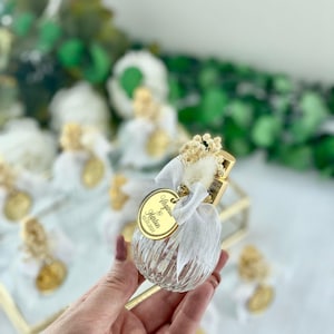 A hand holds a small, clear, ribbed glass perfume bottle with a gold top, white bow, dried flowers, and a gold tag reading "Angelina & Martin 02.10.2023."