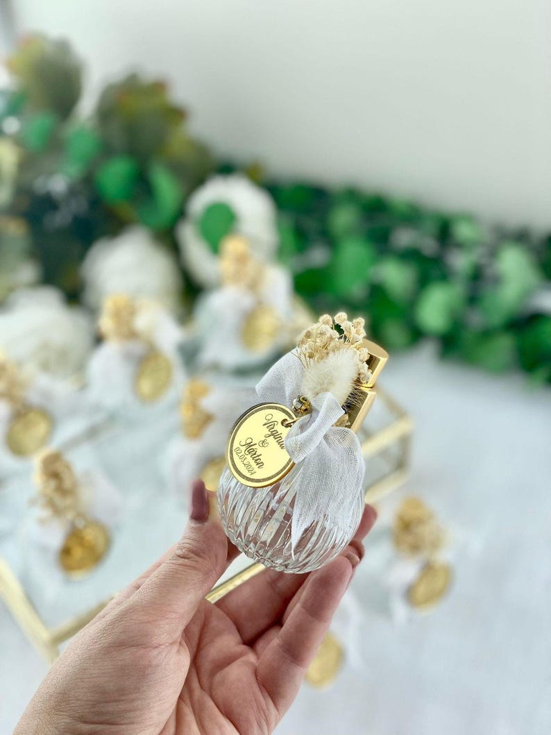 A hand holds a small, clear, ribbed glass perfume bottle with a gold top, white bow, dried flowers, and a gold tag reading "Angelina & Martin 02.10.2023."