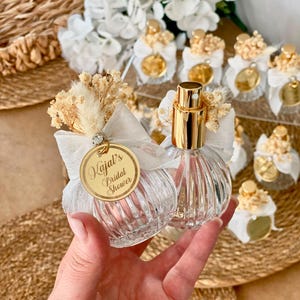 A hand holds two small, clear, ribbed glass perfume bottles with gold tops, white bows, dried flowers, and a gold tag reading "Kajal's Bridal Shower."