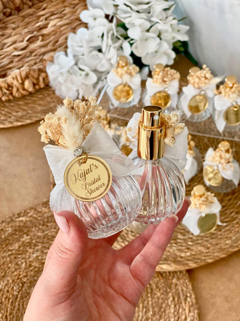 A hand holds two small, clear, ribbed glass perfume bottles with gold tops, white bows, dried flowers, and a gold tag reading "Kajal's Bridal Shower."