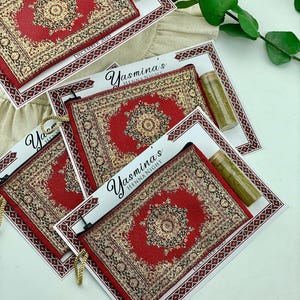 Four red patterned mini wallet favor sets are displayed on a white surface, each with a henna tube and a personalized card.