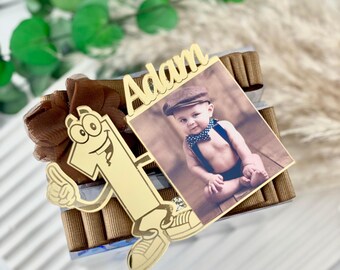 May include: A gold-colored number one cutout with cartoon features and a brown ribbon. A photo of a baby wearing a cap and suspenders is displayed next to the number one. The name "Adam" is written above the photo.