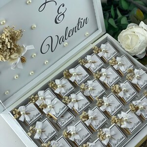 May include: A white box with a gold trim, filled with small white boxes decorated with white bows, pearl beads, and dried flowers. Each box has a label that reads "Leva & Valentin".