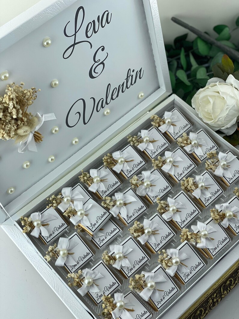 May include: A white box with a gold trim, filled with small white boxes decorated with white bows, pearl beads, and dried flowers. Each box has a label that reads "Leva & Valentin".